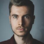 Close-up portrait of a young man with facial hair and a serious expression.